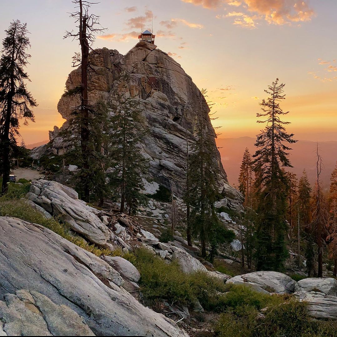 Scenic view of a rocky mountain with a lookout tower on top at sunset, surrounded by trees and a vibrant sky.