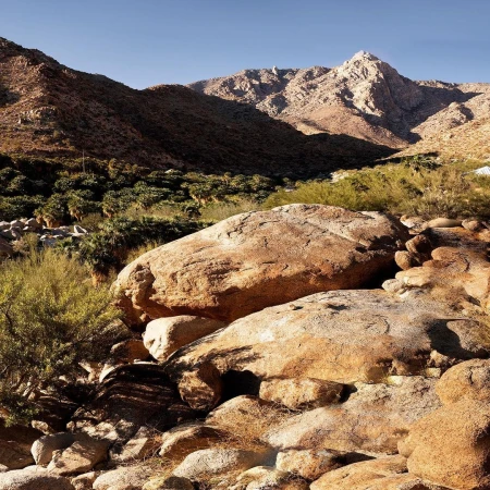 Rocky mountain landscape with large boulders in the foreground, green brush, and a clear blue sky captured by Nina Choi.