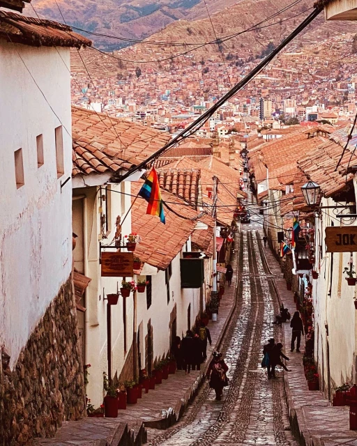 Elevated view down a narrow cobblestone street lined with whitewashed buildings in a city built into a mountainside with colorful flags.