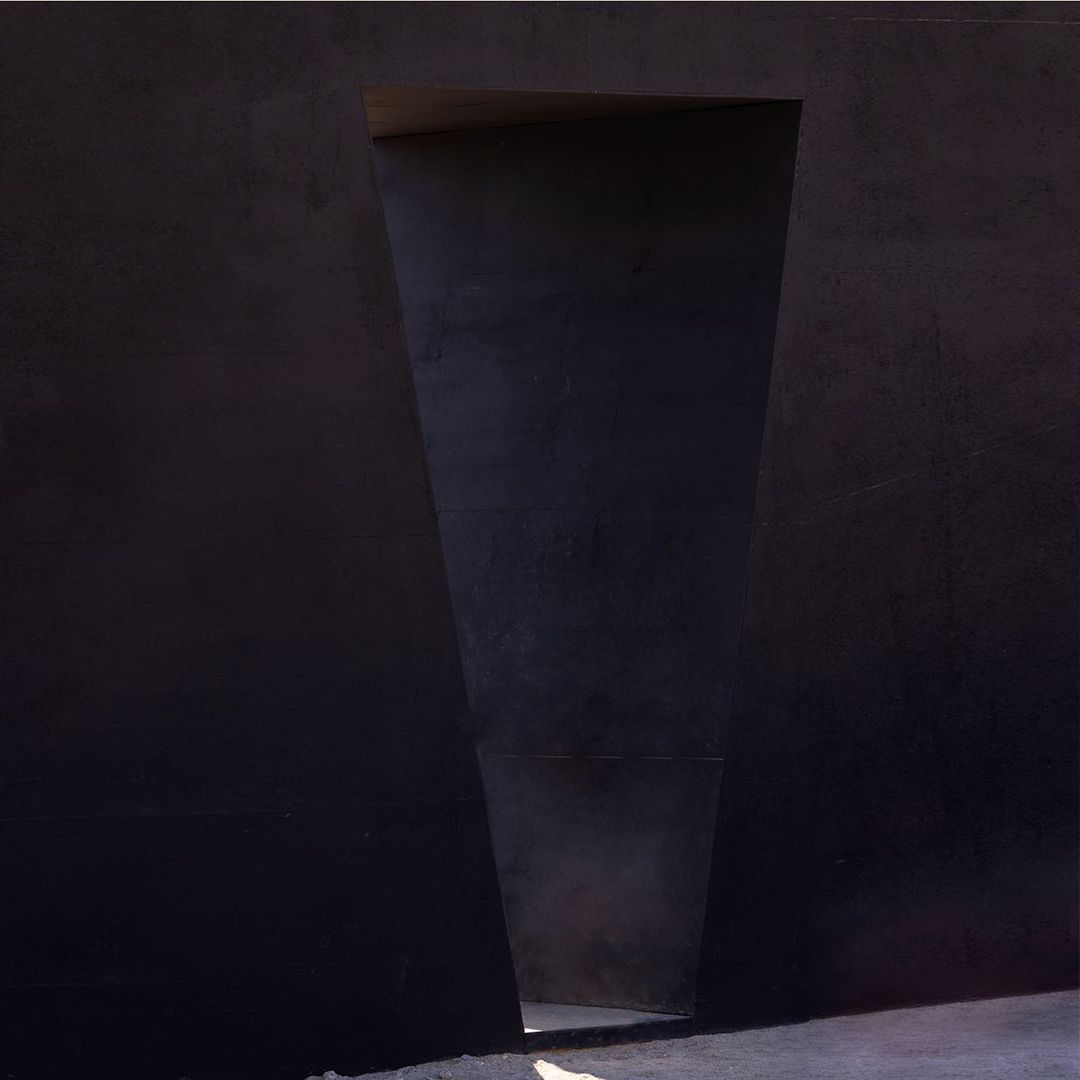 Abstract architectural photograph featuring a dark concrete wall with a trapezoidal opening, revealing light at the end of the passageway.