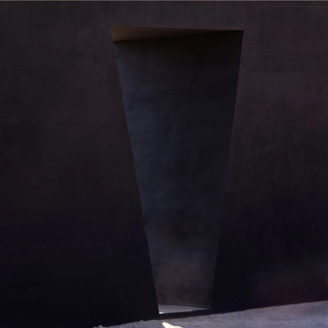 Abstract architectural photograph featuring a dark concrete wall with a trapezoidal opening, revealing light at the end of the passageway.