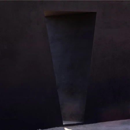 Abstract architectural photograph featuring a dark concrete wall with a trapezoidal opening, revealing light at the end of the passageway.