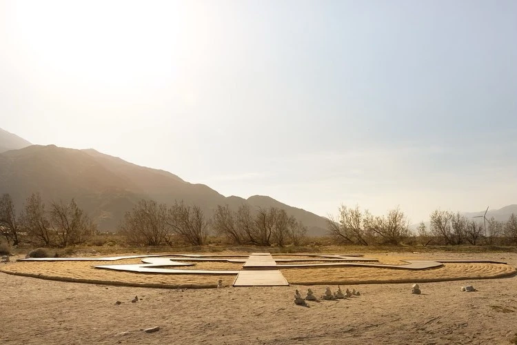 Outdoor desert landscape featuring a sand labyrinth or meditation garden, mountains, wind turbines, and a clear sky.