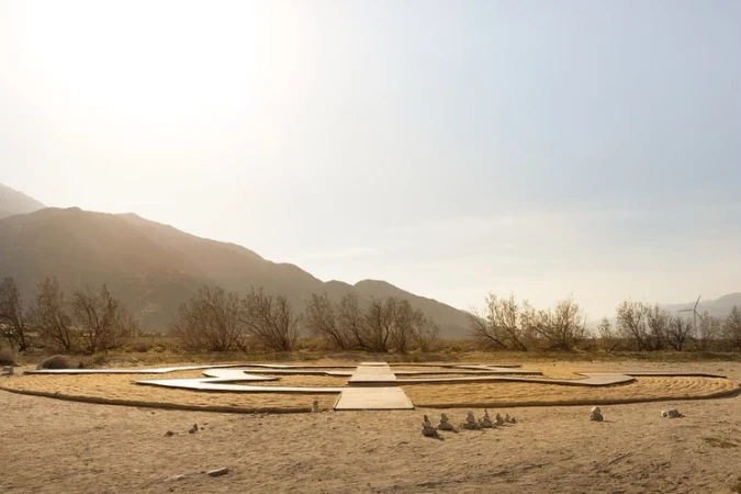 Outdoor desert landscape featuring a sand labyrinth or meditation garden, mountains, wind turbines, and a clear sky.