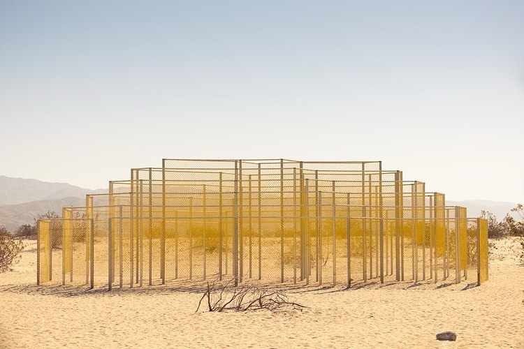 Minimalist outdoor shot of a yellow wireframe structure in a desert landscape under a clear blue sky, creating a unique geometric pattern.