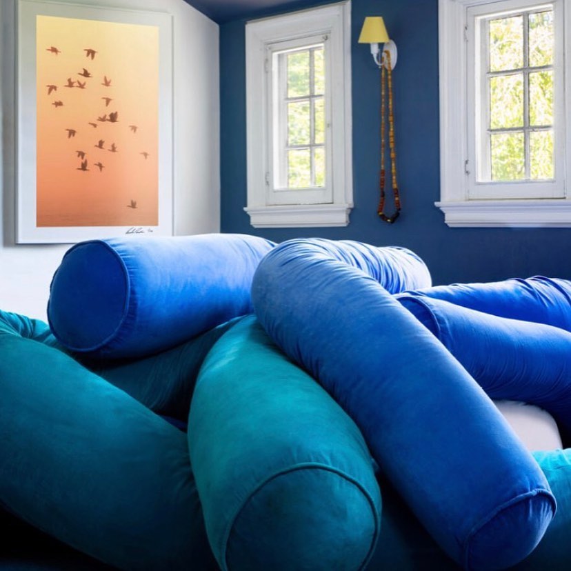 Bolster pillows in shades of teal and blue are stacked on a couch inside a living room with a nature-themed print and white-framed windows.