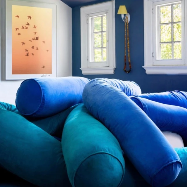 Close-up interior view of vibrant blue bolster pillows piled on furniture against a blue wall with two white windows
