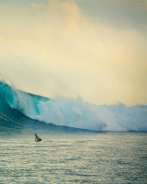 Surfer on a surfboard riding a massive ocean wave with white crest and spray under a pale sky