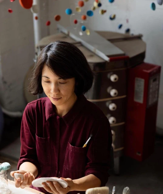 Asian woman wearing a burgundy button-down shirt working in a pottery studio with a kiln in the background