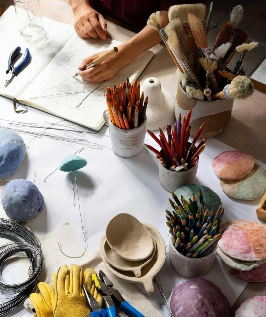 Overhead view of an artist drawing in a sketchbook surrounded by colored pencils, tools, ceramics, and sketches on a desk.