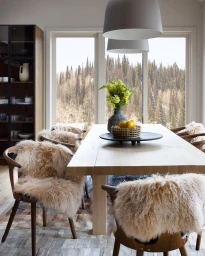 Wooden dining table with fur-covered chairs in an interior space with a large window framing a pine forest view.