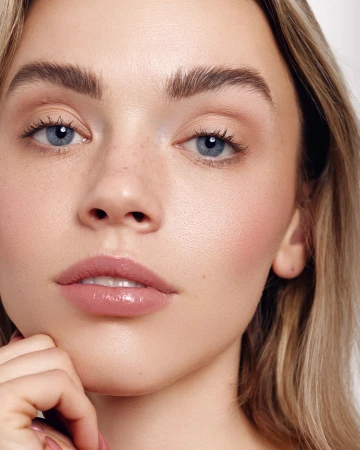 Extreme close-up portrait of a woman with blonde hair, blue eyes, and freckles, her hand resting near her chin against a white background