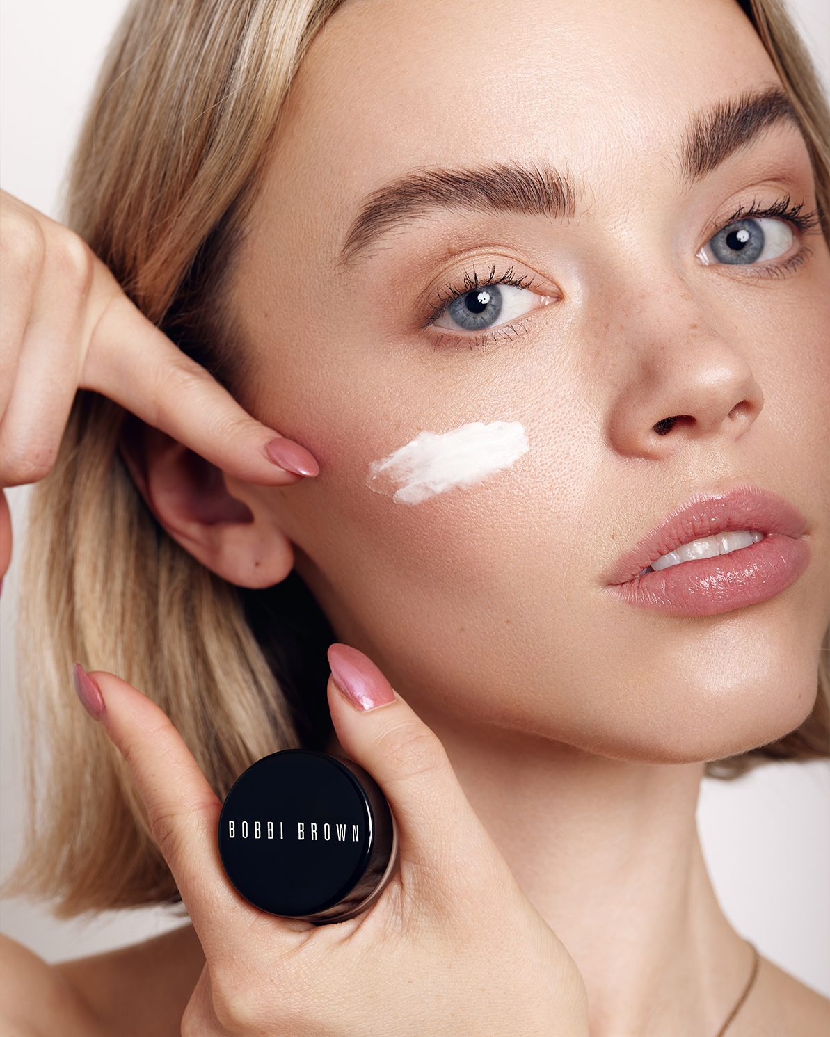 Close up of a blonde woman applying white cream to her cheek with a small jar of Bobbi Brown skincare product in her hand.