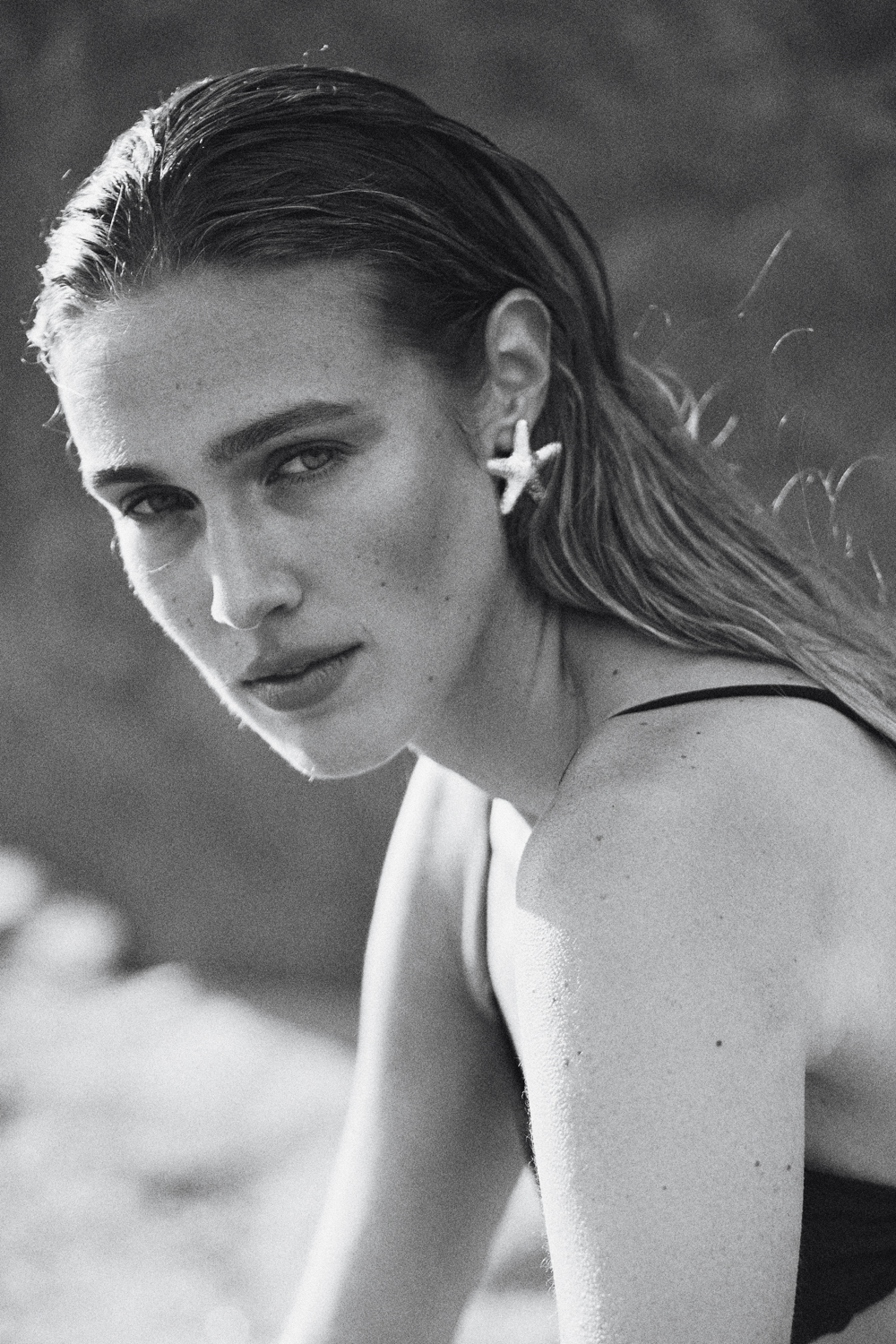Portrait of a woman with slicked-back hair wearing a starfish earring, looking directly at the camera in a black and white photograph.