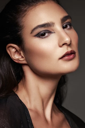 Close-up portrait of a woman with bold graphic eyeliner, arched eyebrows, and dark lips, looking directly at the camera.