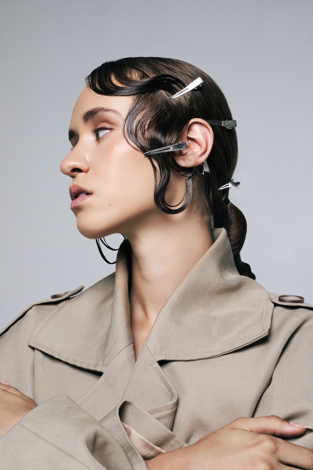 A profile portrait of a person with finger wave hair styled with multiple silver metal hair clips, wearing a beige trench coat.