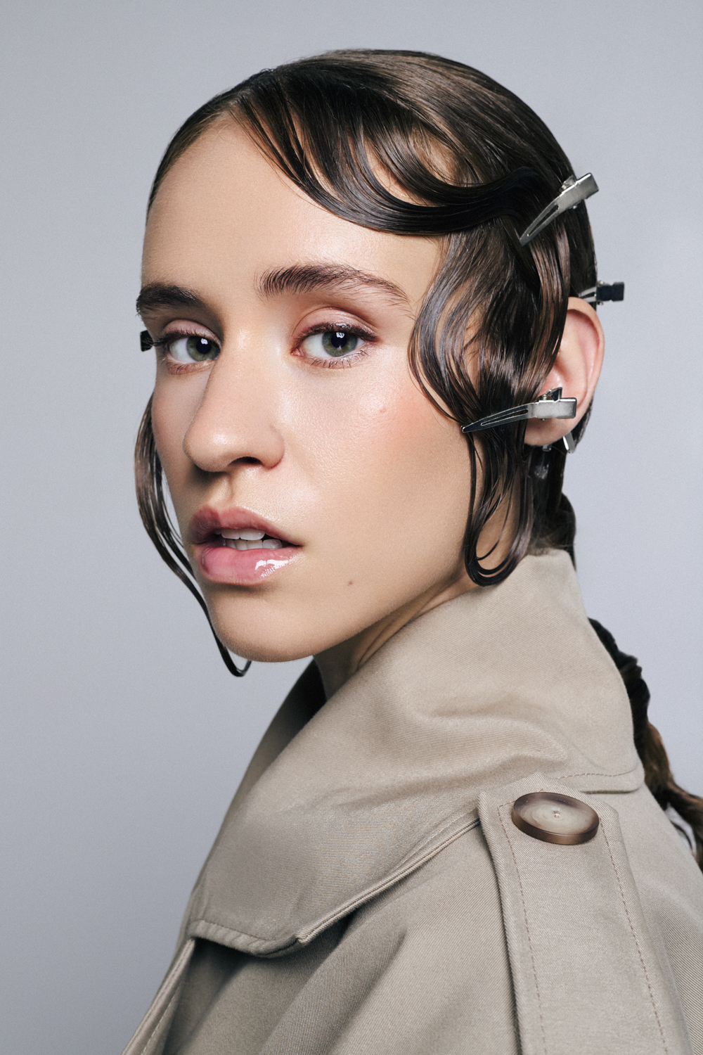 Close-up portrait of a woman with wet-look hair styled with silver metal hair clips, wearing a neutral beige trench coat.
