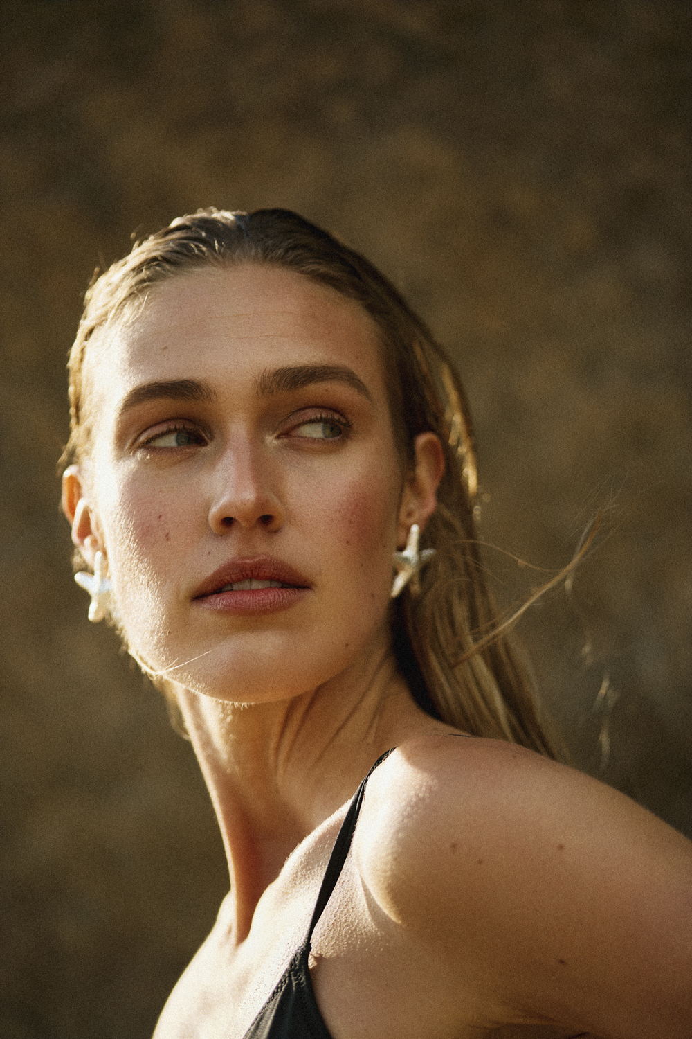 Close-up portrait of a woman with wet hair wearing starfish-shaped earrings, looking away from the camera in warm natural light.
