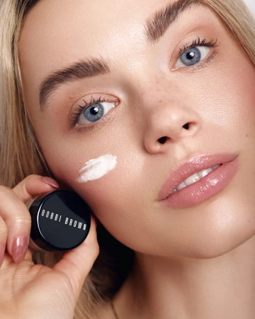 Close-up of a blonde woman with blue eyes holding a small black jar of Bobbi Brown cream, a dab of white cream on her cheek.