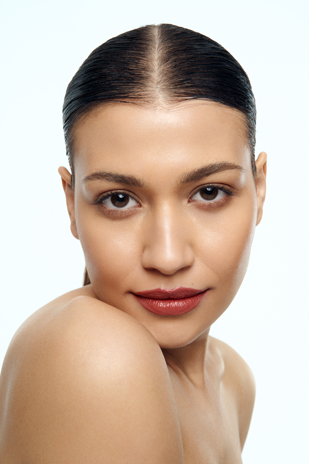 Close-up beauty portrait of a woman with slicked-back dark hair, bare shoulders, and glossy lips against a white background.