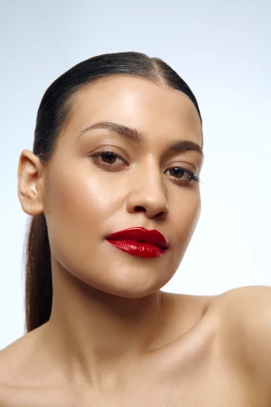 Close-up of a woman with sleek dark hair in a ponytail and vibrant glossy red lipstick against a plain light gray studio background