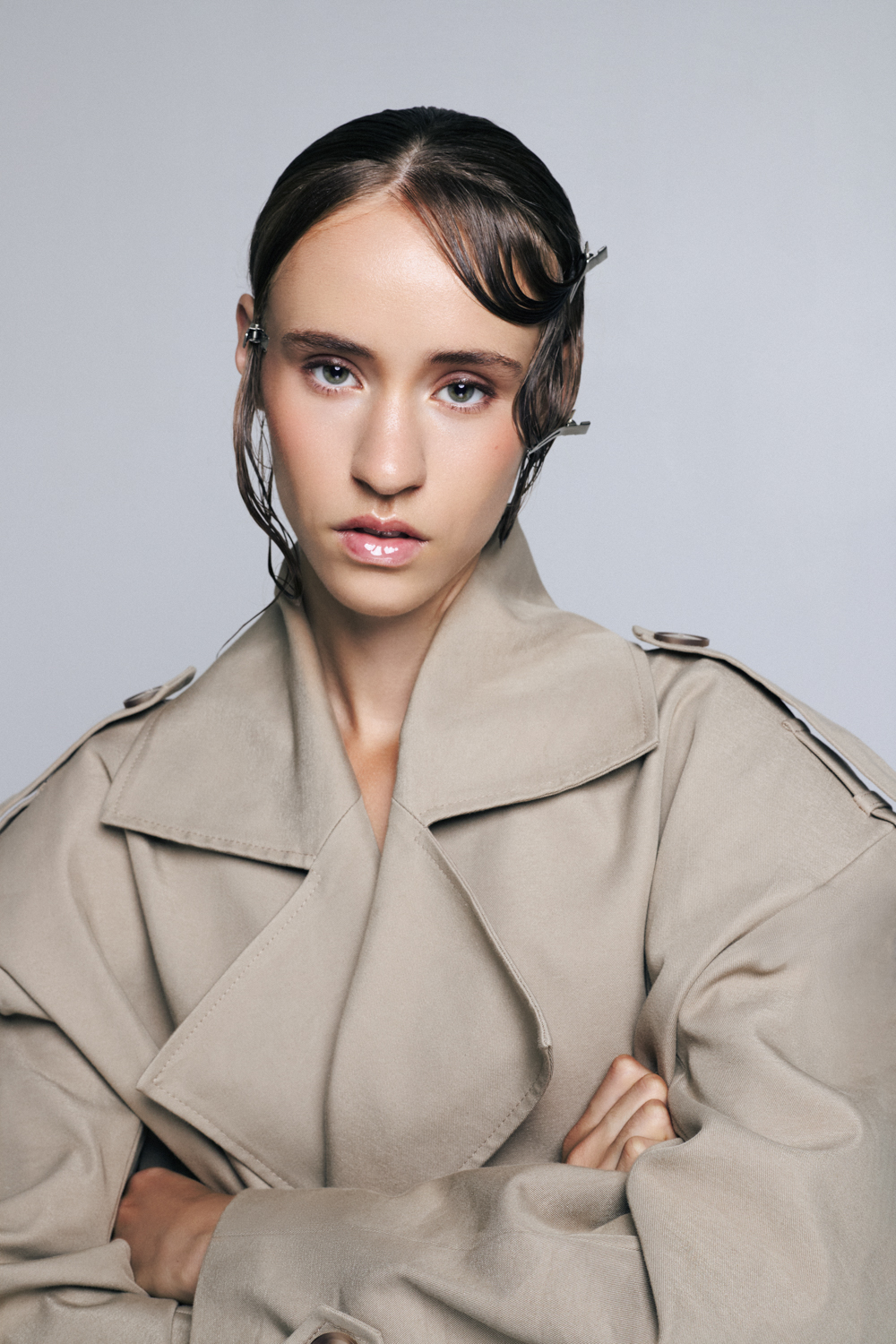 A woman with dark hair pinned back with metal clips wearing a beige trench coat stands with arms crossed against a light gray background.