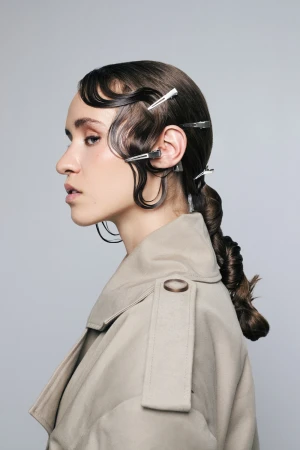Profile view of a woman with wet-look hair styled in waves and secured with silver metal hair clips, wearing a tan trench coat.