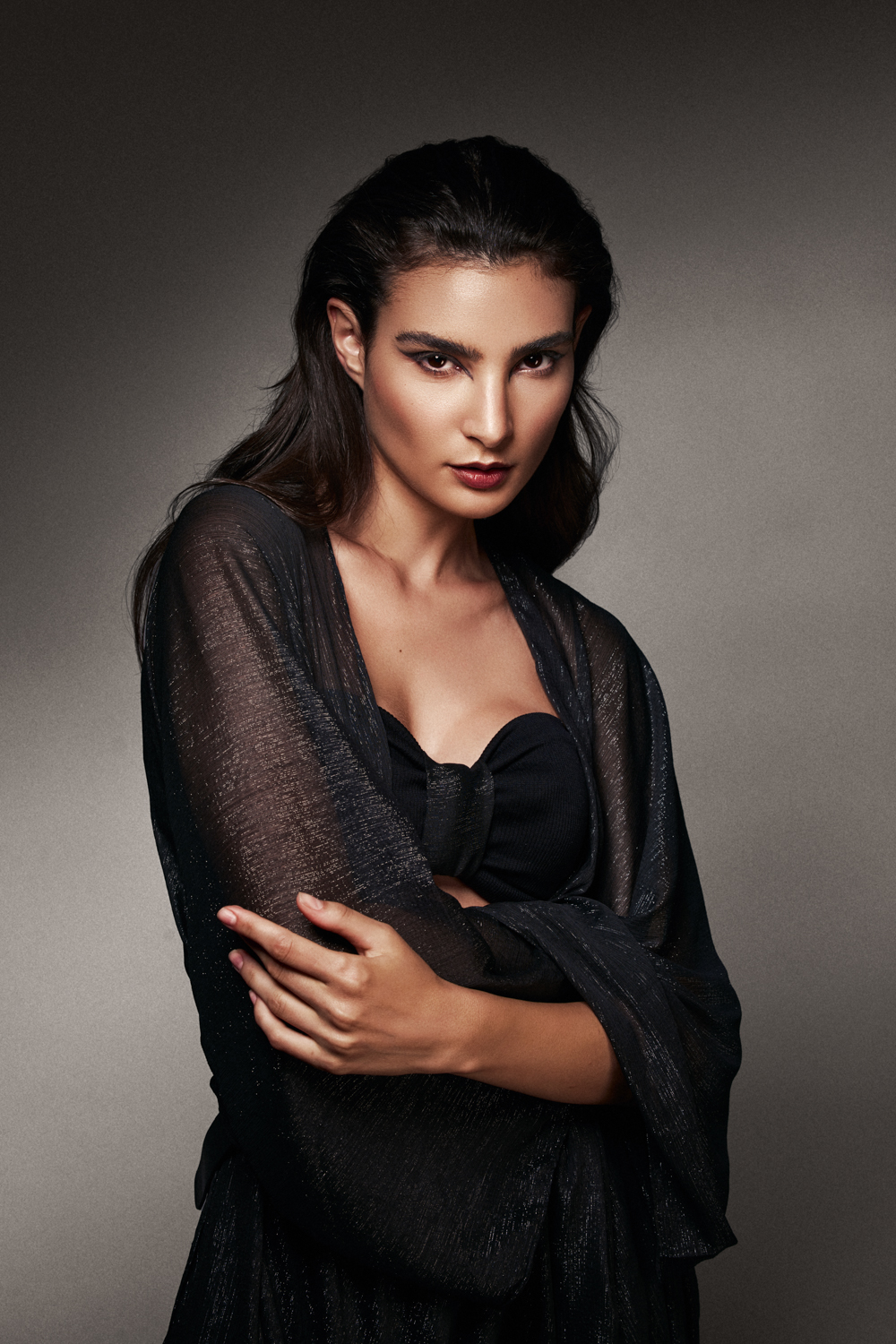 Close-up portrait of a woman with dark hair and dark lipstick, wearing a black sheer shawl over a structured bodice against a grey background.
