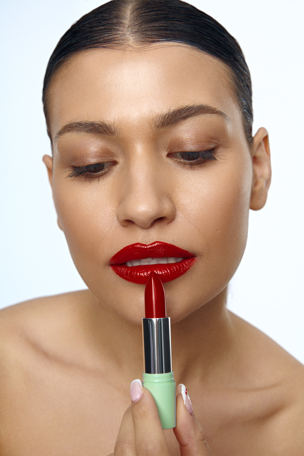 Close-up of a person with dark hair slicked back applying a vibrant red lipstick to their glossy lips against a clean white background.