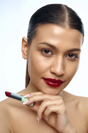 Close-up portrait of a woman with slicked-back dark hair, wearing glossy deep red lipstick and holding a matching lipstick tube to her chin.