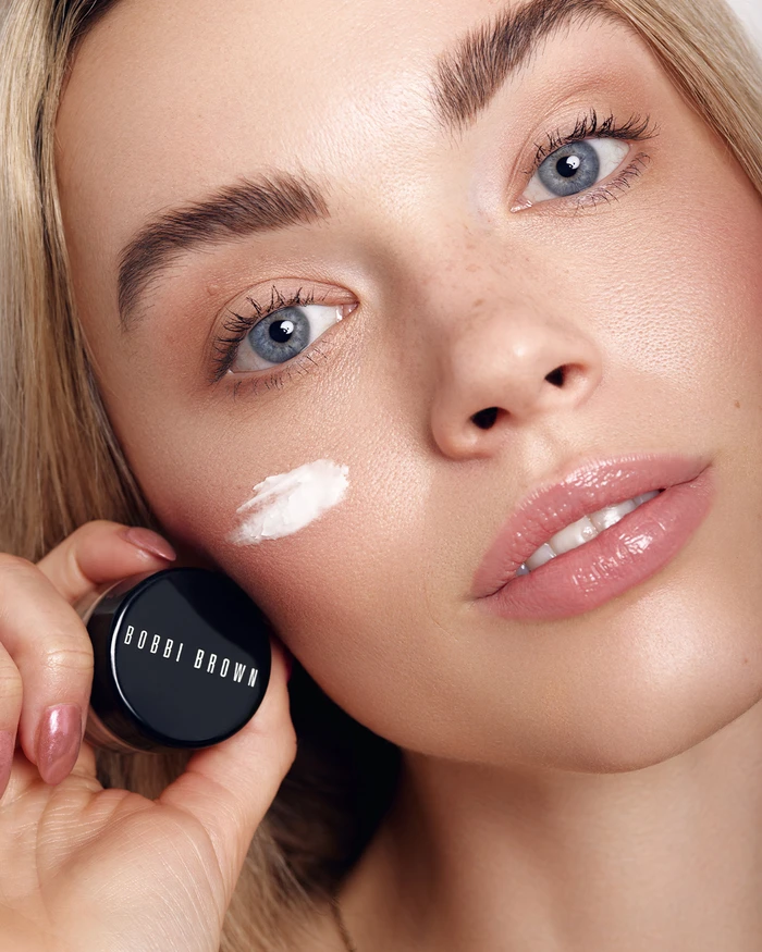 Close-up beauty portrait of a woman holding a Bobbi Brown jar with white cream on her cheek and glossy nude lipstick.