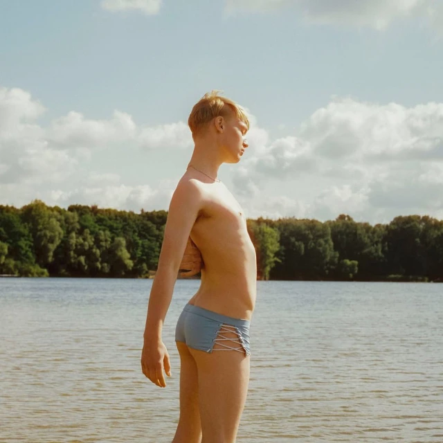 Young adult stands in swimwear on a beach, looking out over lake water towards a background of trees under a cloudy sky.