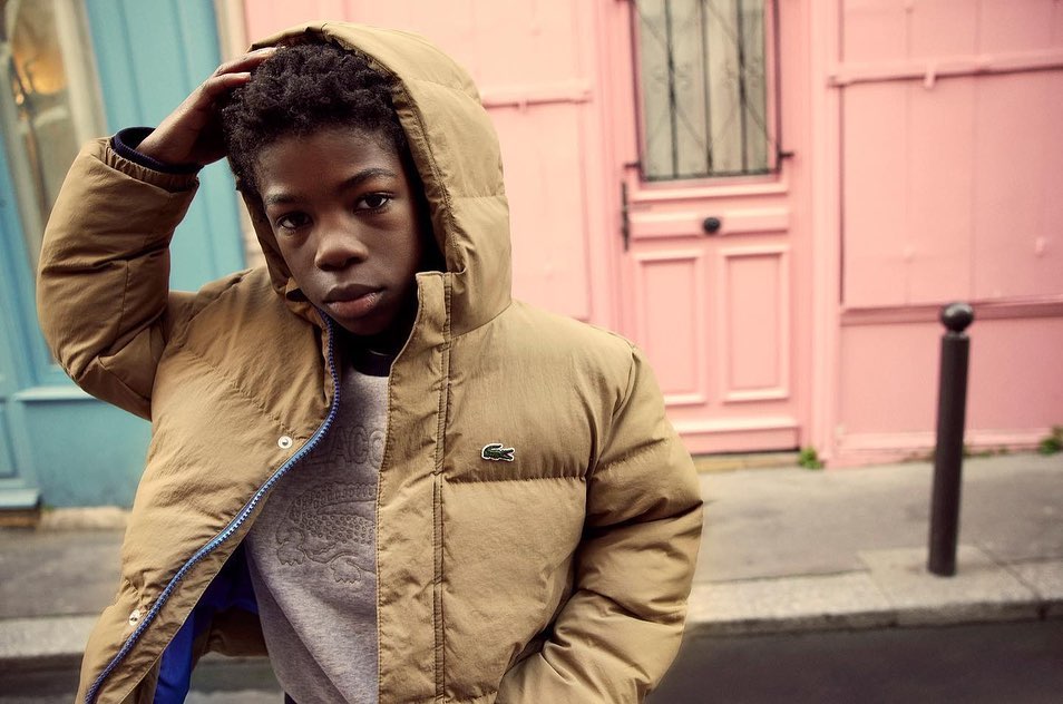 Young person in puffer jacket poses in front of a pink building with blue accents. Street style fashion shot in an urban environment.