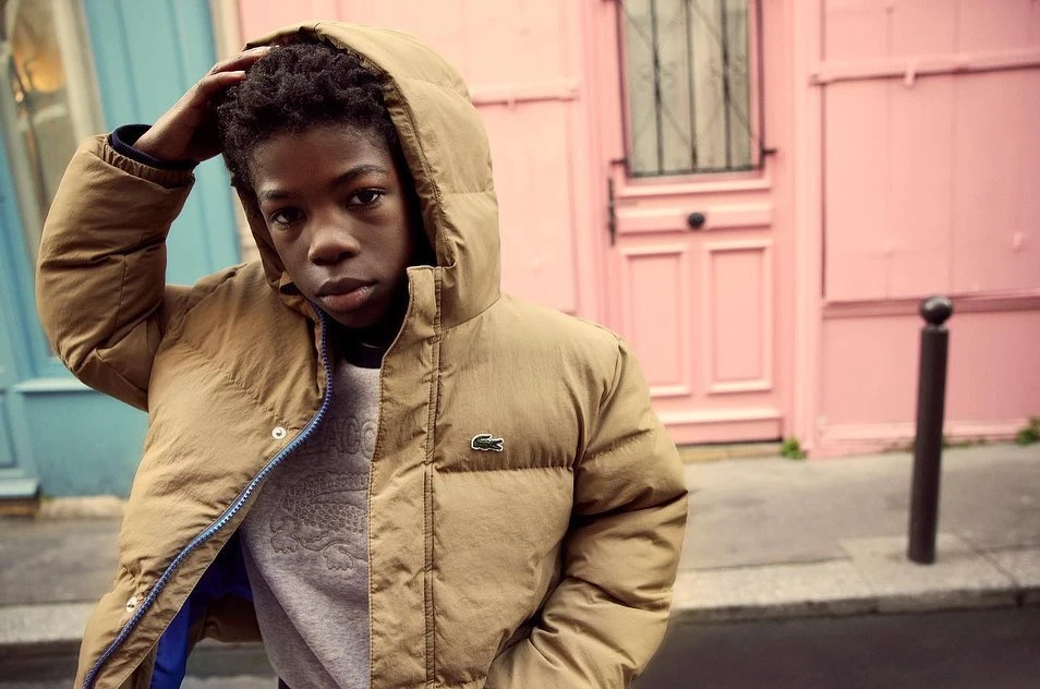 Young person in puffer jacket poses in front of a pink building with blue accents. Street style fashion shot in an urban environment.