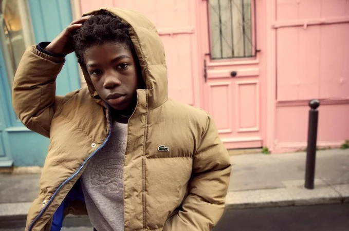 Young person in puffer jacket poses in front of a pink building with blue accents. Street style fashion shot in an urban environment.
