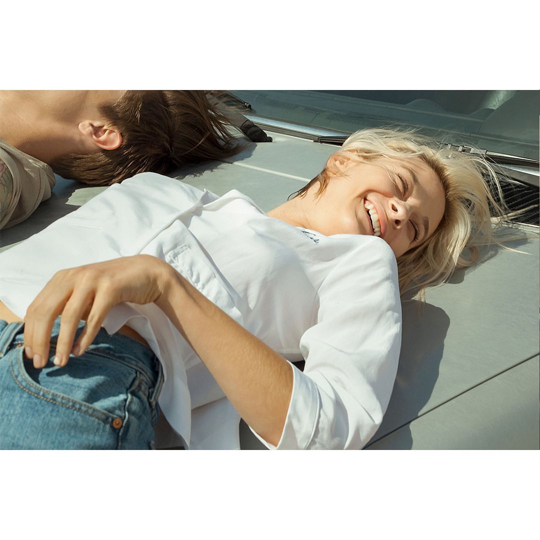 Two fair-skinned people lying on a gray surface outdoors, one smiling in a white shirt and jeans under bright sunlight. 