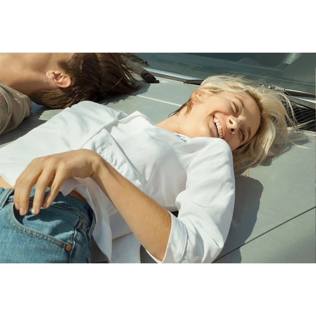 Two fair-skinned people lying on a gray surface outdoors, one smiling in a white shirt and jeans under bright sunlight. 