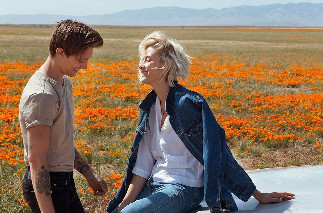Couple smiling in orange poppy field, woman in blue jean jacket. Mountains in distance, medium shot, daytime, outdoor setting.