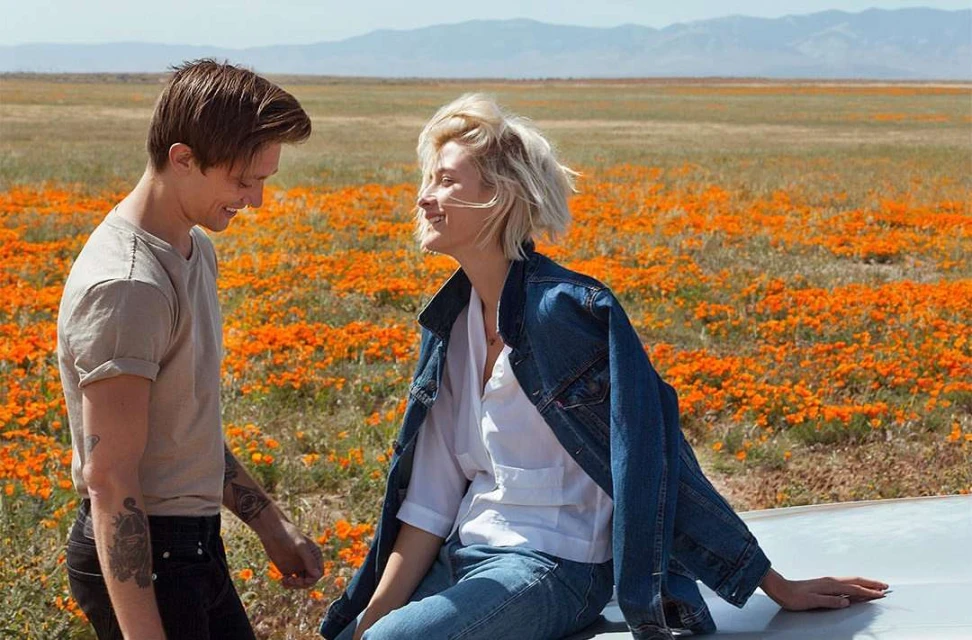 Couple smiling in orange poppy field, woman in blue jean jacket. Mountains in distance, medium shot, daytime, outdoor setting.