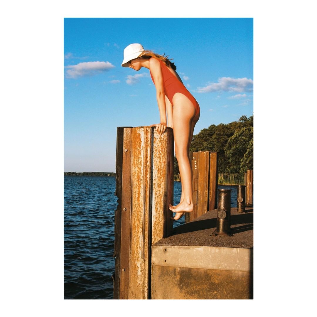 A young woman in a red swimsuit and white bucket hat stands on a wooden pier overlooking the water on a sunny day.