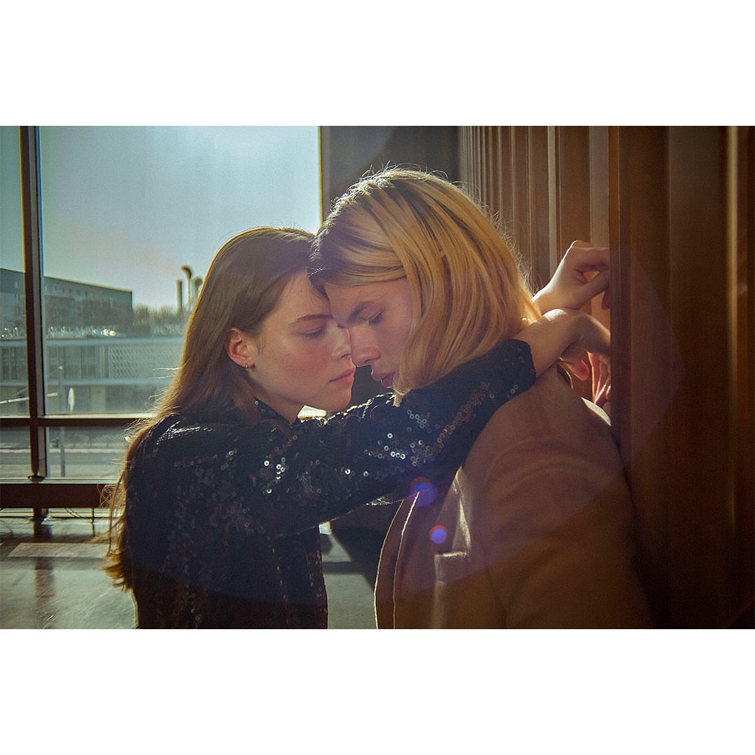 A young couple embraces indoors near a large window, casting a warm light on their faces and highlighting their intimate moment.