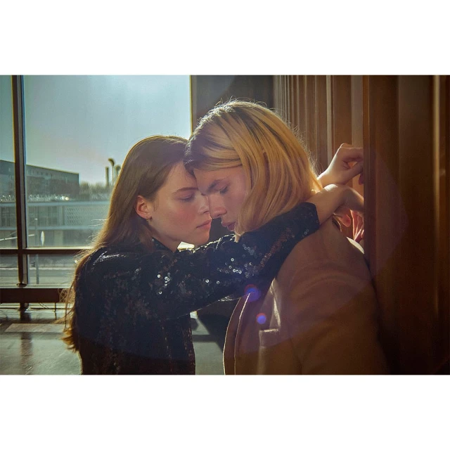 A young couple embraces indoors near a large window, casting a warm light on their faces and highlighting their intimate moment.