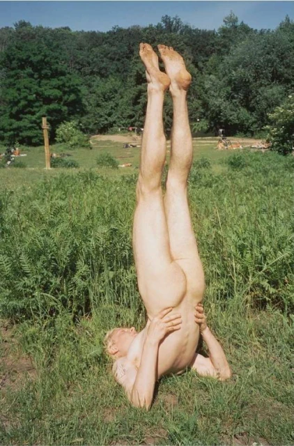 Nude adult in the supported shoulderstand yoga pose on a grassy field with trees and sunbathers visible in the background on a sunny day.