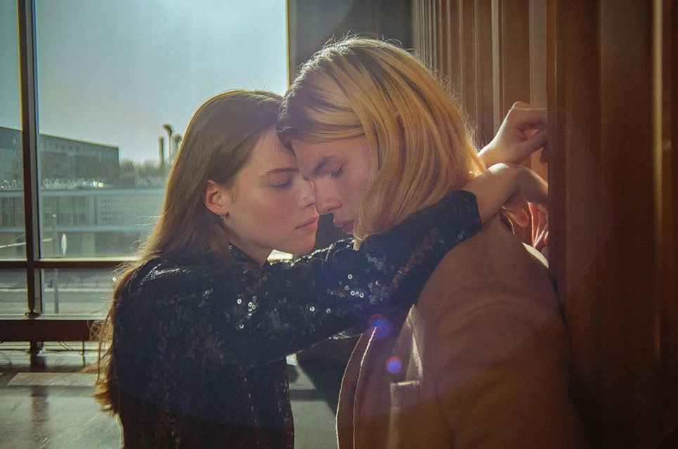 A close-up of a young couple embracing indoors by a wooden railing, with an out-of-focus building visible through the window in the background.