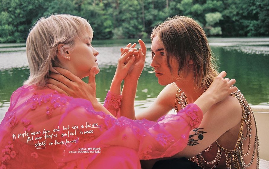 Two people holding hands in a lake setting; woman with short blonde hair and a pink dress, and person with long brown hair.