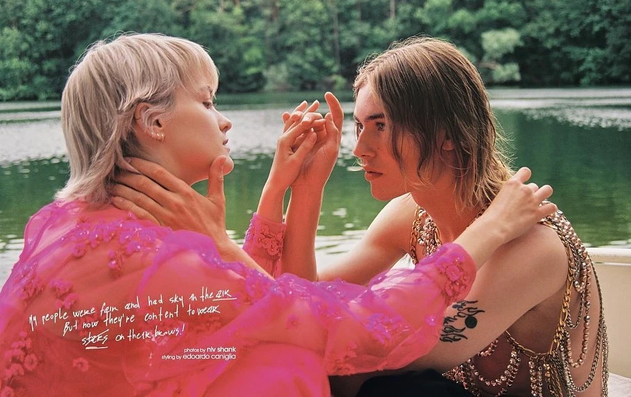 Two people holding hands in a lake setting; woman with short blonde hair and a pink dress, and person with long brown hair.