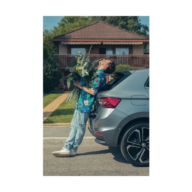 Man in a floral shirt leans against a gray car trunk while holding large bouquet. Residential house and tree-lined street in background.