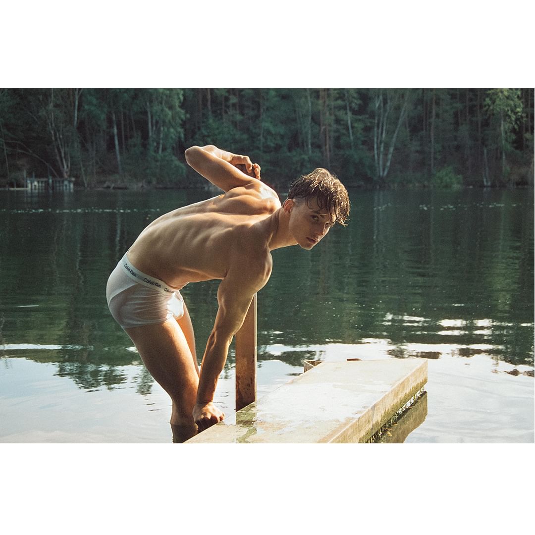 A shirtless young man in briefs stands on a wooden platform in a lake, with a forest in the background, creating a natural, outdoor scene.