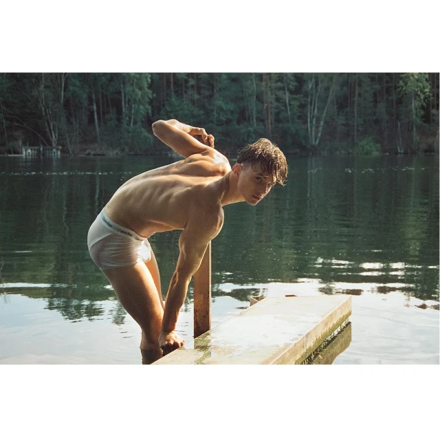 A shirtless young man in briefs stands on a wooden platform in a lake, with a forest in the background, creating a natural, outdoor scene.