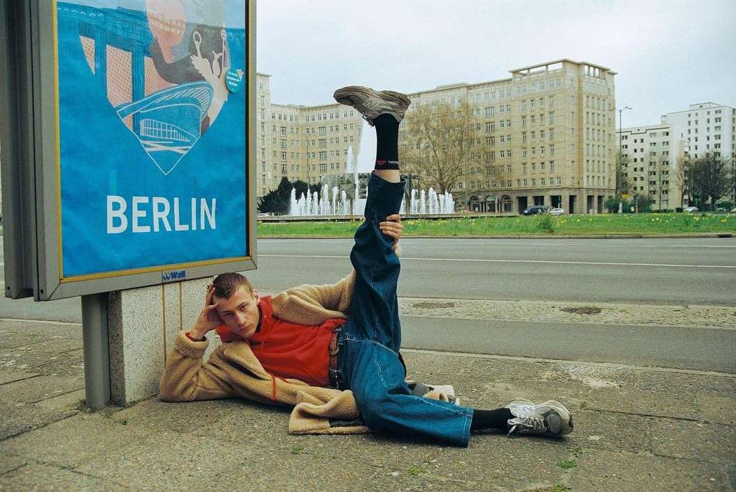 Man in red shirt and jeans poses on ground next to Berlin tourism billboard, buildings and fountain in the background on overcast day.